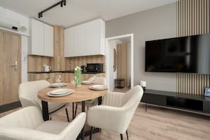 A modern kitchen with white cabinets, a wooden accent wall, a dining table with chairs, a TV, and a small side table.