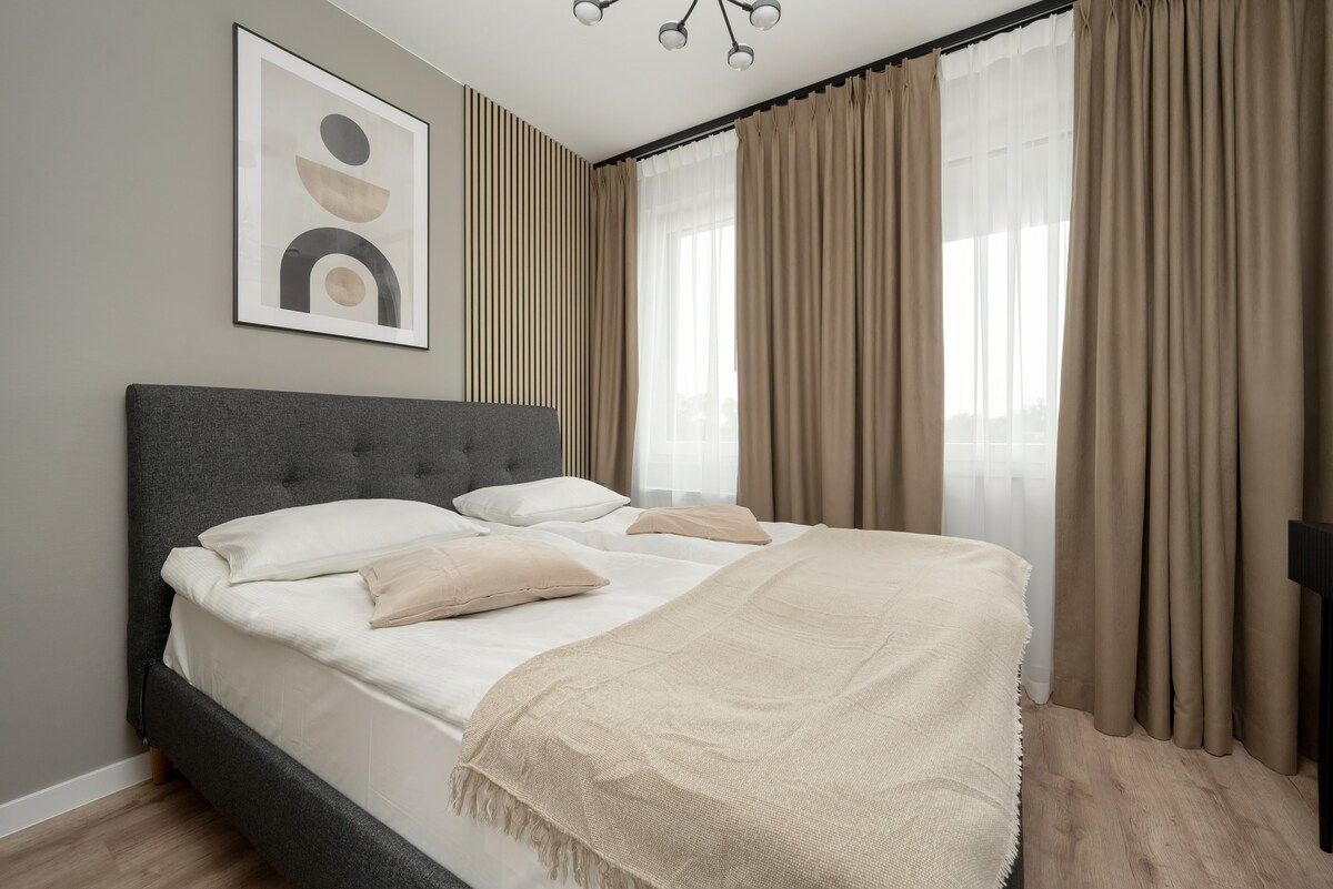 Un dormitorio con cama, cabecera, un cuadro enmarcado en la pared y cortinas.