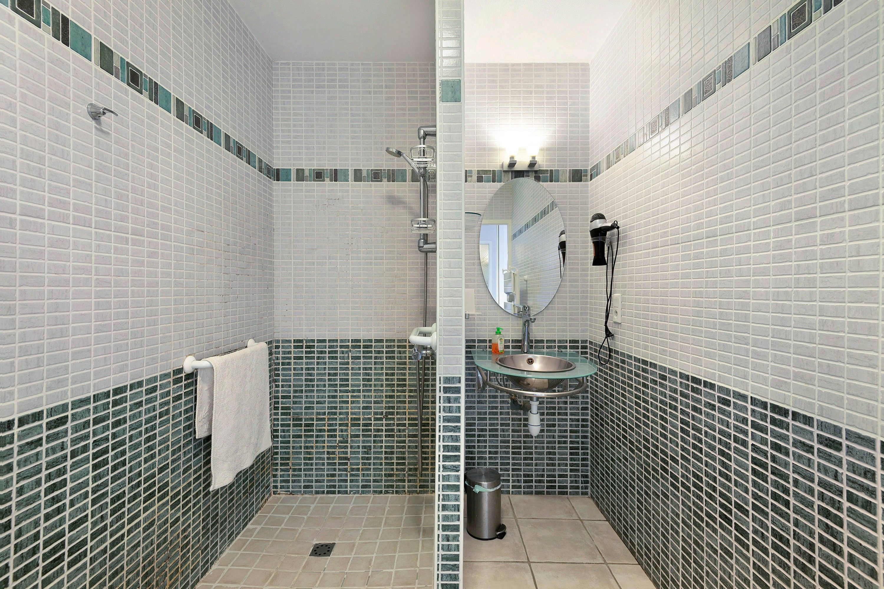 Une salle de bain avec des murs recouverts de carreaux blancs, un lavabo en verre, une douchette à main, un porte-serviettes et une poubelle.