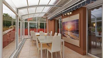 Een overdekt terras met een eettafel en stoelen, een groot raam met uitzicht op de tuin, en een kunstwerk aan de muur.