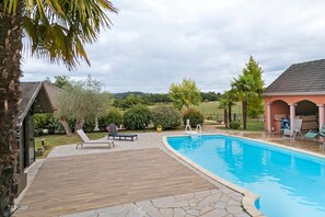 Una piscina con muelle de madera, sillas de descanso y un área techada.
