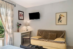 A room with a bed, a sofa, a desk with a chair, a TV mounted on the wall, and a window with curtains.