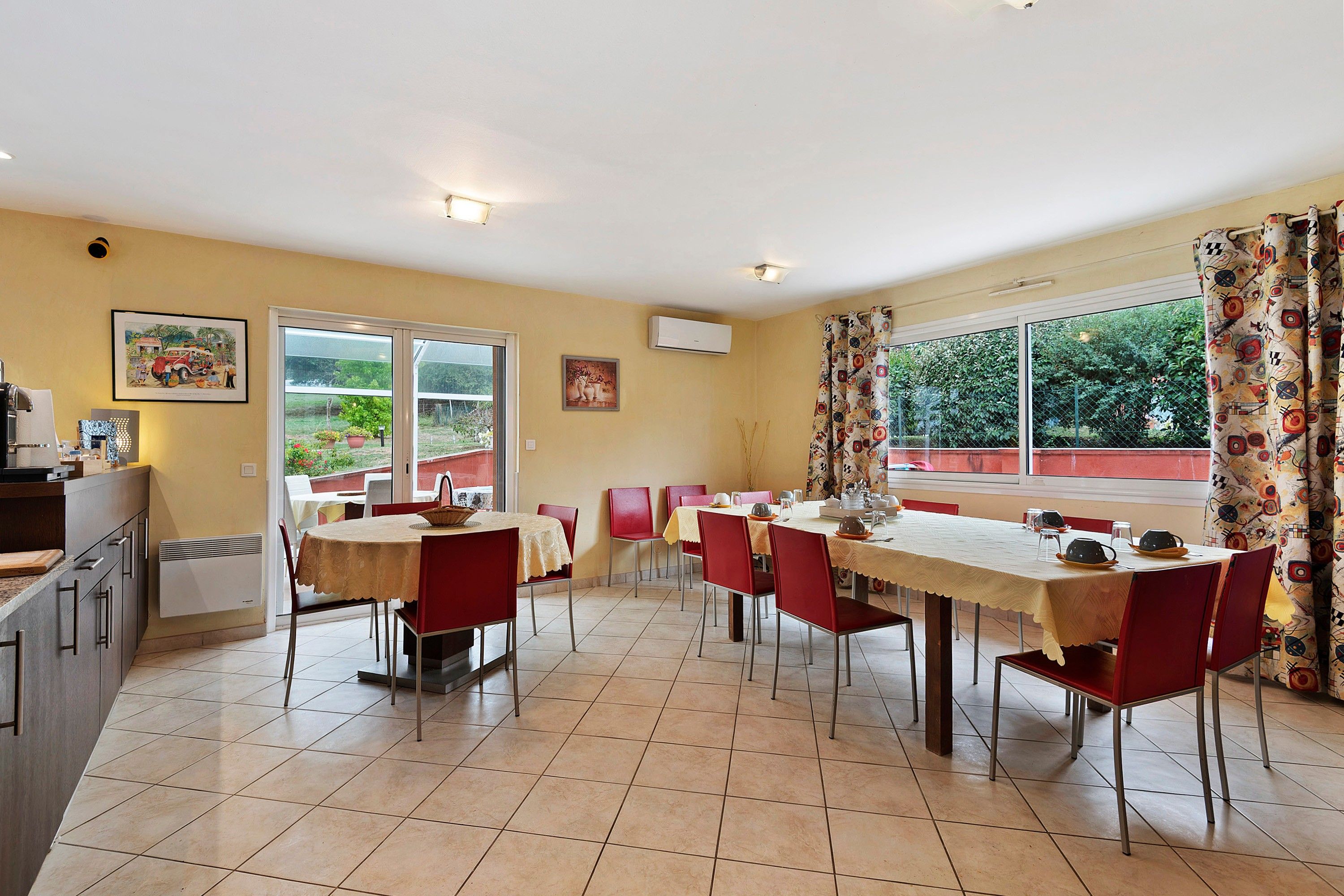 Un coin repas avec une table dressée pour quatre personnes, des chaises rouges et une vue sur la verdure à l’extérieur.