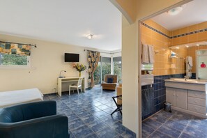 A hotel room with a desk, chair, sofa, and a bathroom with a sink and mirror.