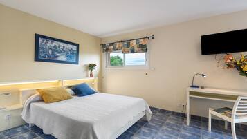 Une chambre à coucher comprenant un lit, un bureau, une chaise, une télévision, une fenêtre avec des rideaux, un tableau accroché au mur et un vase avec des fleurs.