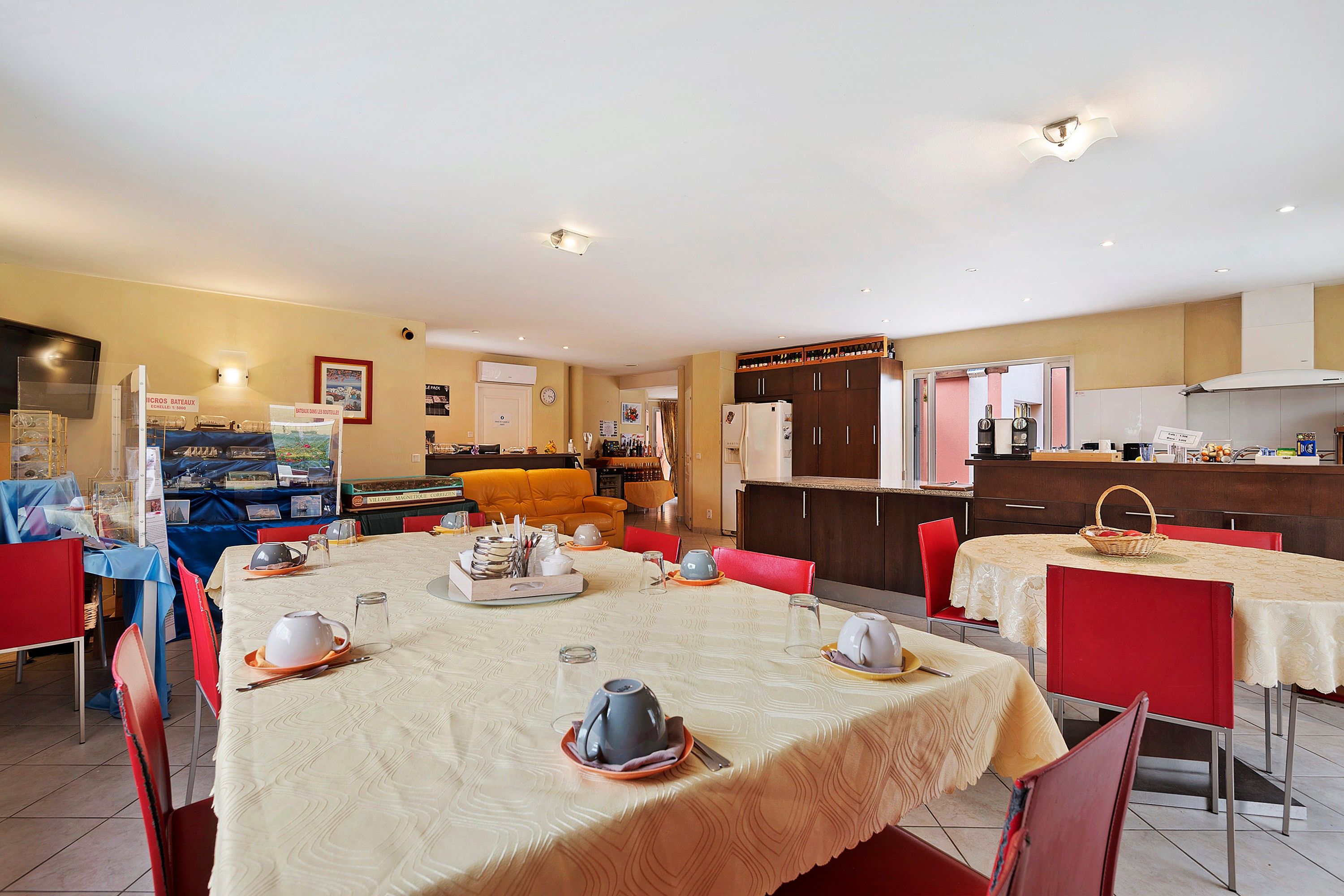 Un coin repas avec des tables dressées pour le repas, une cuisine en arrière-plan et un espace de vie avec un canapé.