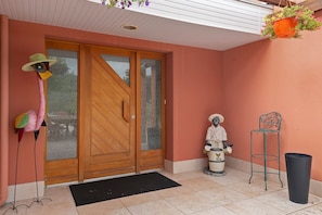 Una puerta de madera con un panel de vidrio, un felpudo negro en la entrada, una silla de metal, un cubo de basura y una maceta colgante.