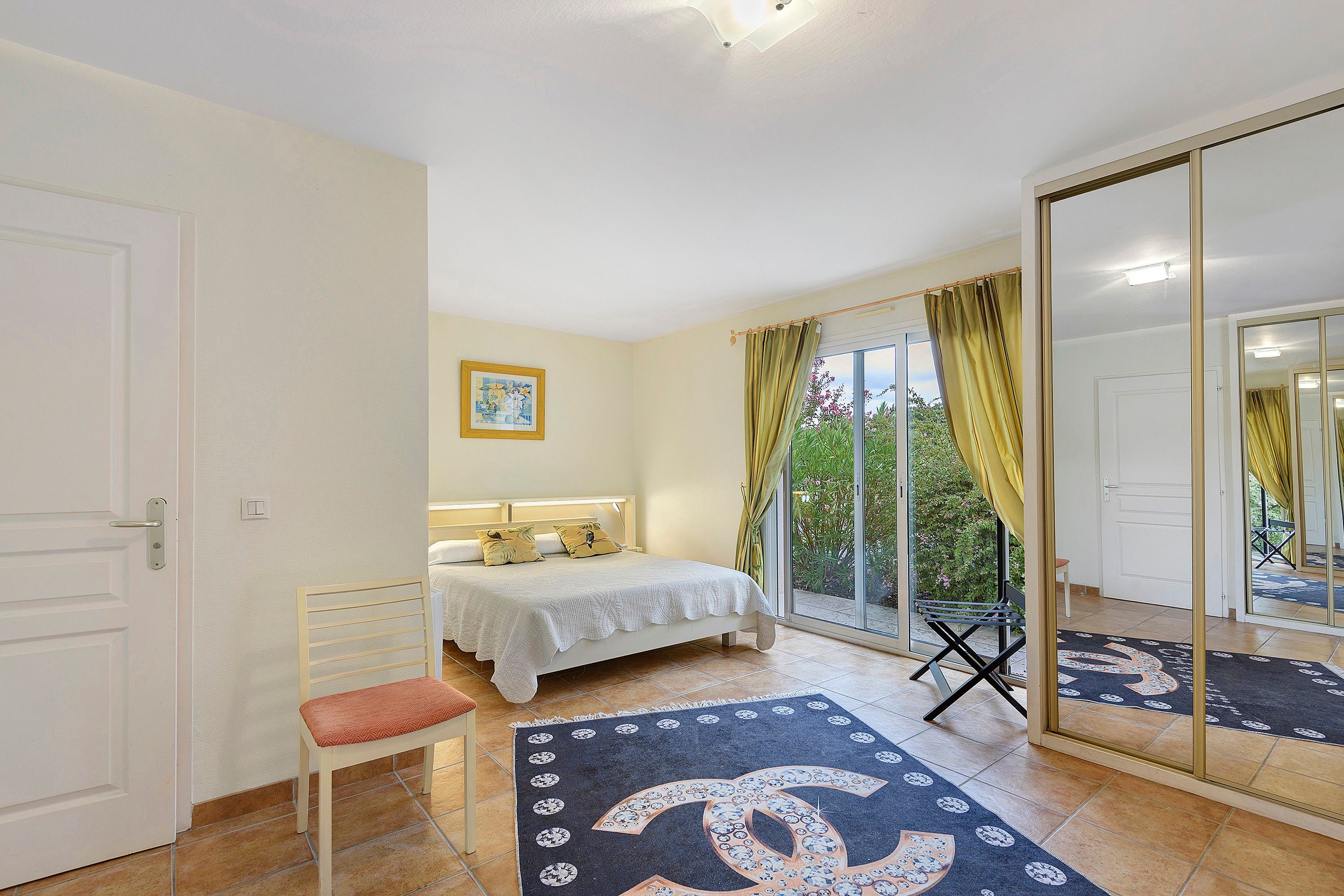 Une chambre à coucher avec un lit, une chaise, un grand miroir et une vue sur la verdure à l’extérieur.