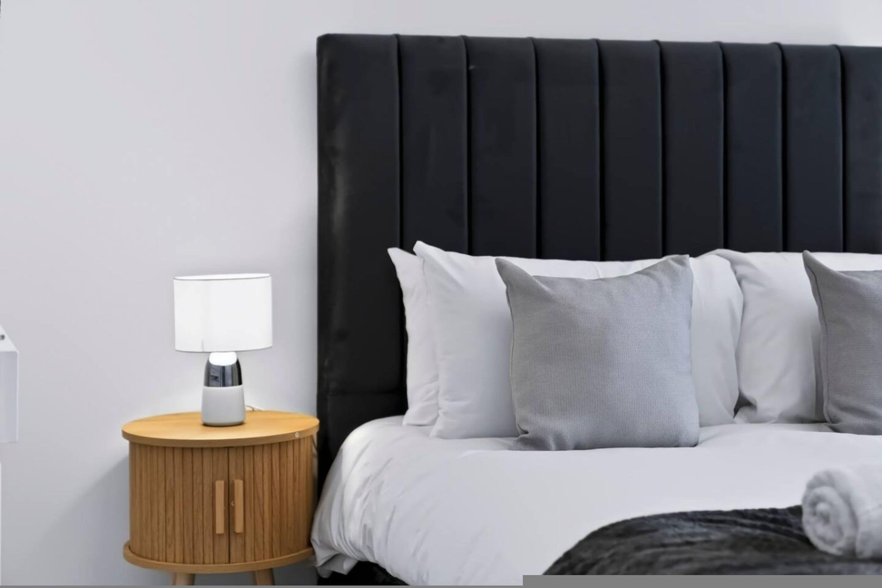 A bed with a black headboard, white bedding, and a wooden bedside table with a lamp.