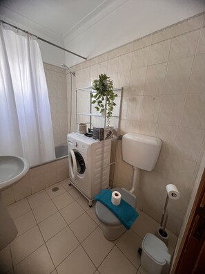 A compact bathroom with a washing machine, toilet, and a shelf with a plant. - Cantinho de Mar Holiday Home (Monte Gordo)