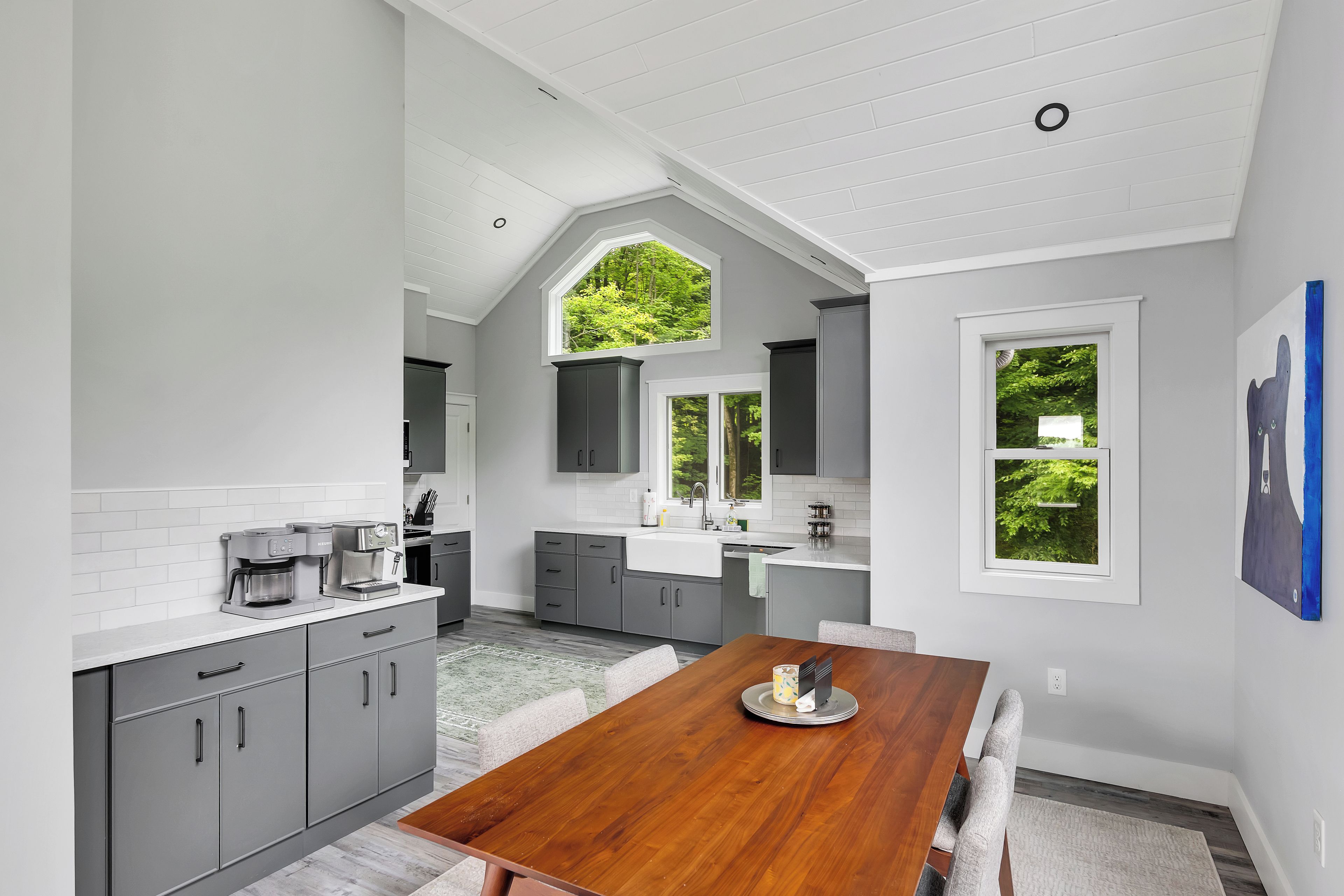 Una cocina moderna con armarios grises, una mesa de comedor de madera y vistas a zonas verdes por las ventanas.