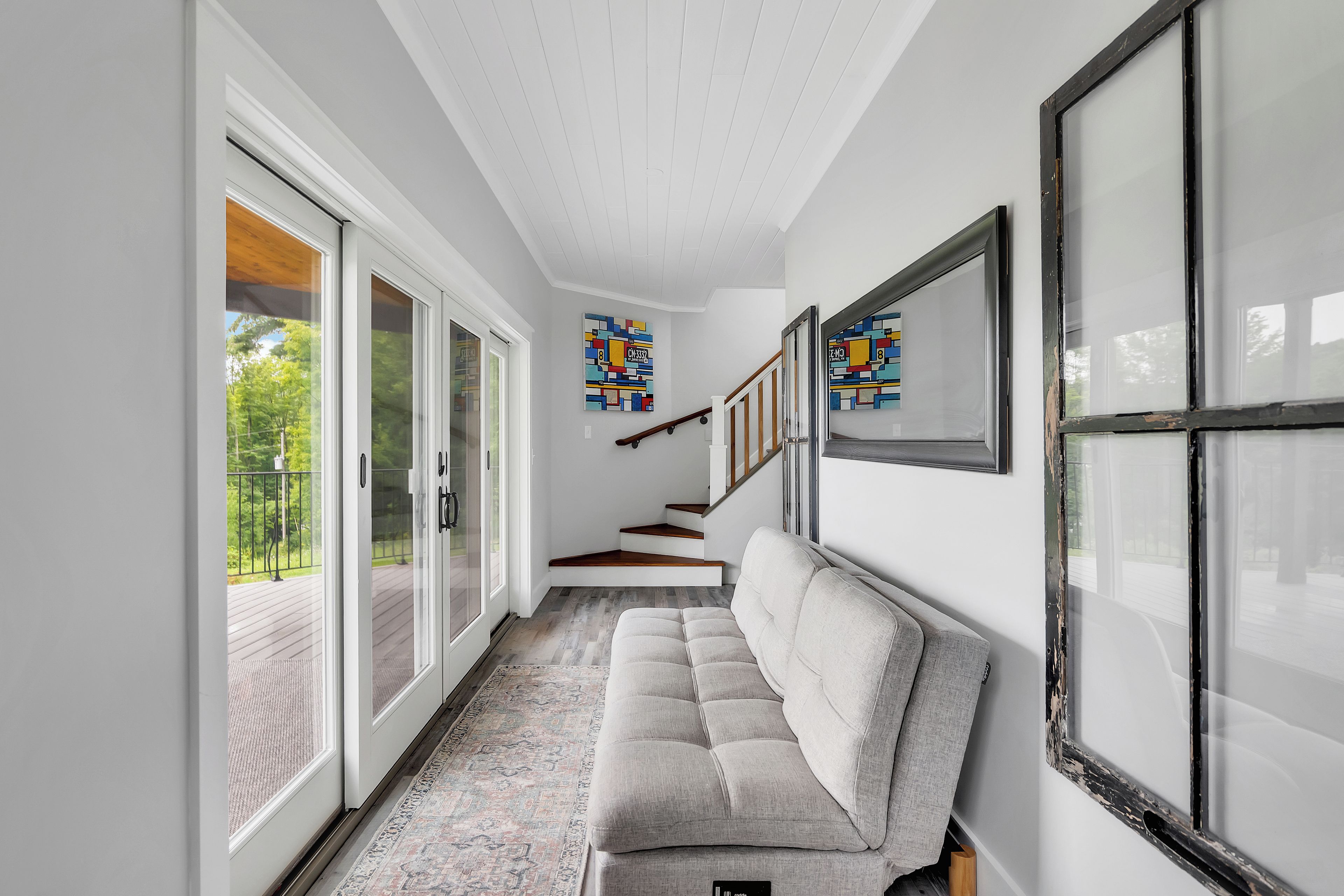 Un pasillo con un sofá gris, una escalera y vistas al exterior con vegetación.