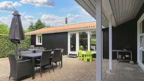Eine Terrasse im Freien mit Korbmöbeln, einem Tisch und Stühlen, einem überdachten Bereich mit Grill und Blick auf ein Haus mit Ziegeldach.