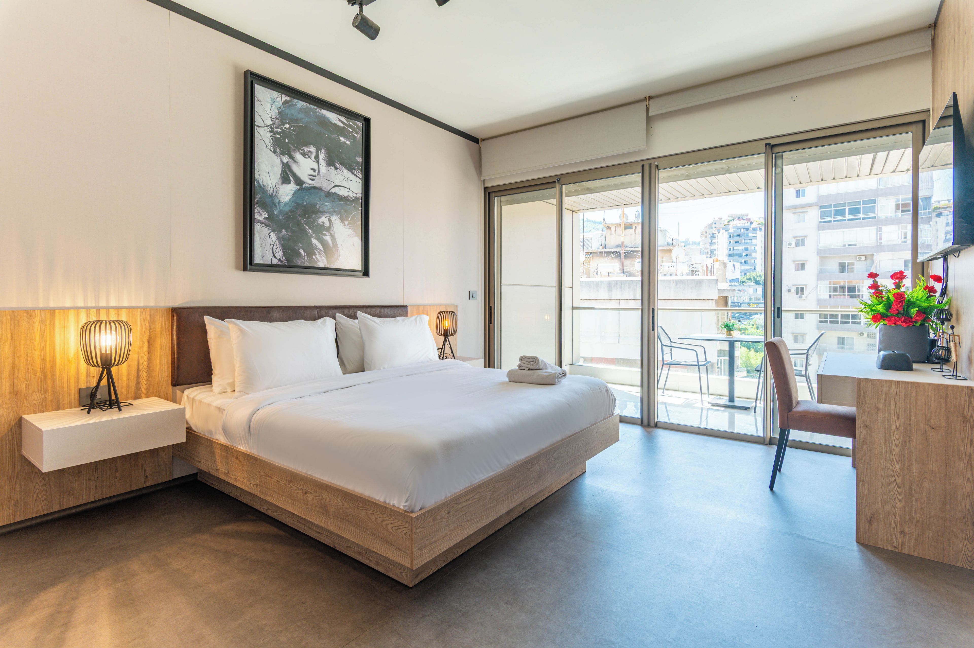 Una habitación de hotel moderna con una cama grande, una mesita de noche con lámpara, un cuadro en la pared y un balcón con vistas a la ciudad.