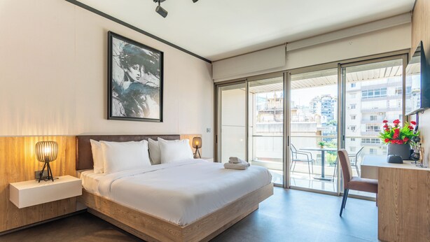 A modern hotel room with a large bed, a bedside table with a lamp, a framed artwork on the wall, and a balcony with a view of the city. - Elara Studio & Balcony in Jounieh (Jounieh)