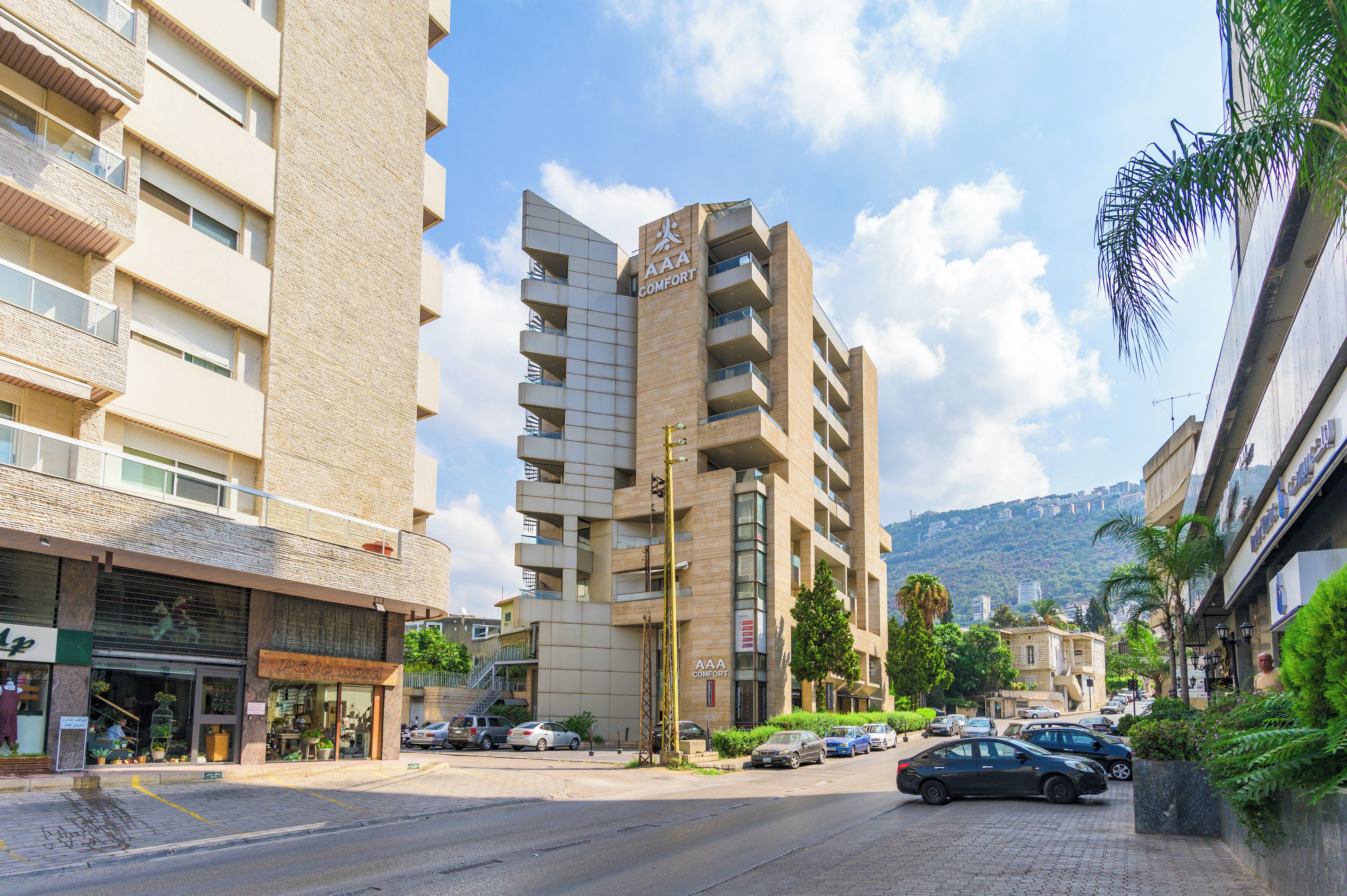 Un edificio de varios pisos con balcones, un letrero que dice 'AAA COMFORT' y una calle con coches estacionados y vegetación.