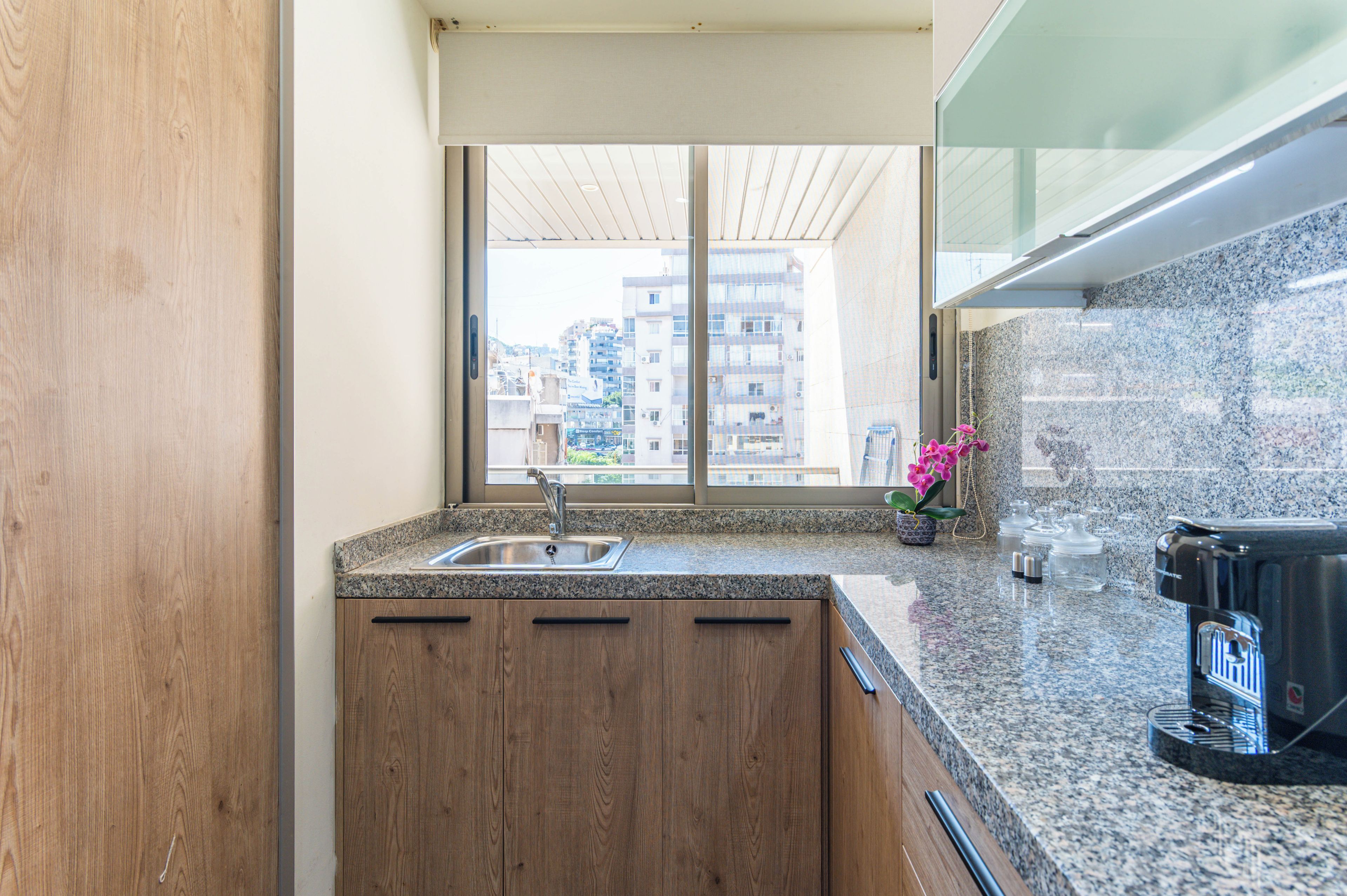 Eine moderne Küche mit einer Granit-Arbeitsplatte, Holzschränken, einer Edelstahl-Spüle und Blick auf eine Stadtlandschaft durch ein großes Fenster.