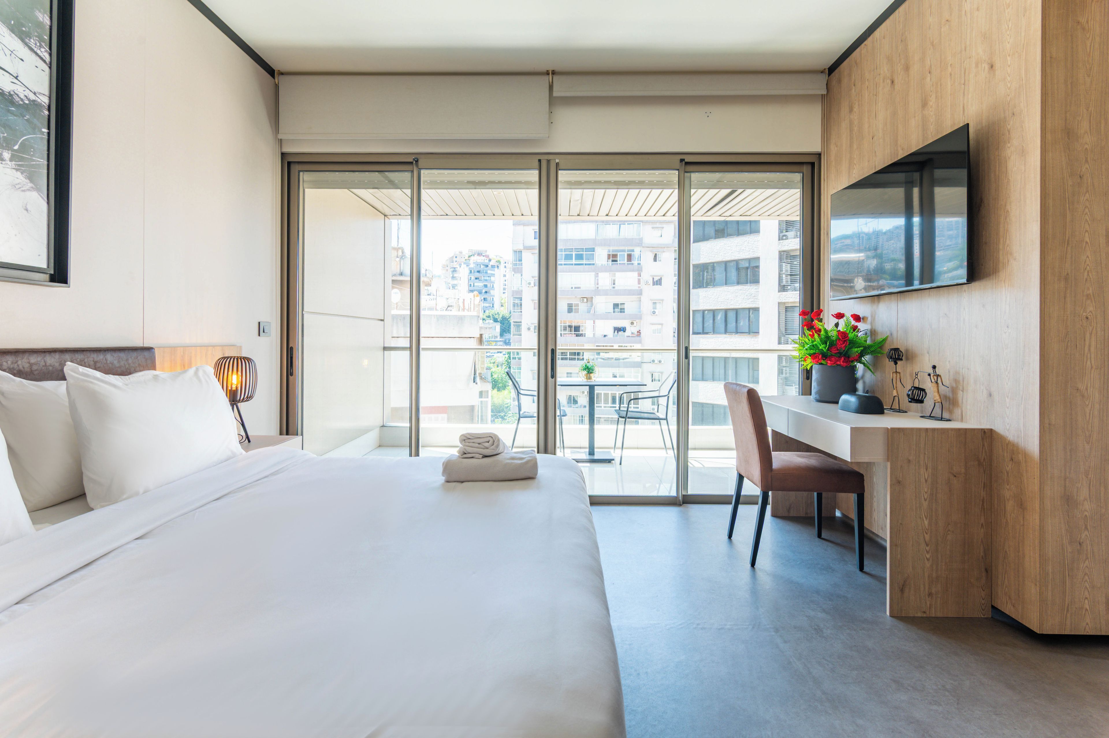 Habitación de hotel con cama, escritorio, silla, televisor y balcón con vista a edificios.