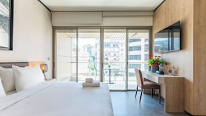 A hotel room with a bed, a desk, a chair, a TV, and a balcony with a view of buildings. - Elara Studio & Balcony in Jounieh (Jounieh)