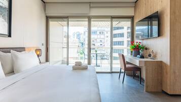 Habitación de hotel con cama, escritorio, silla, televisor y balcón con vista a edificios.
