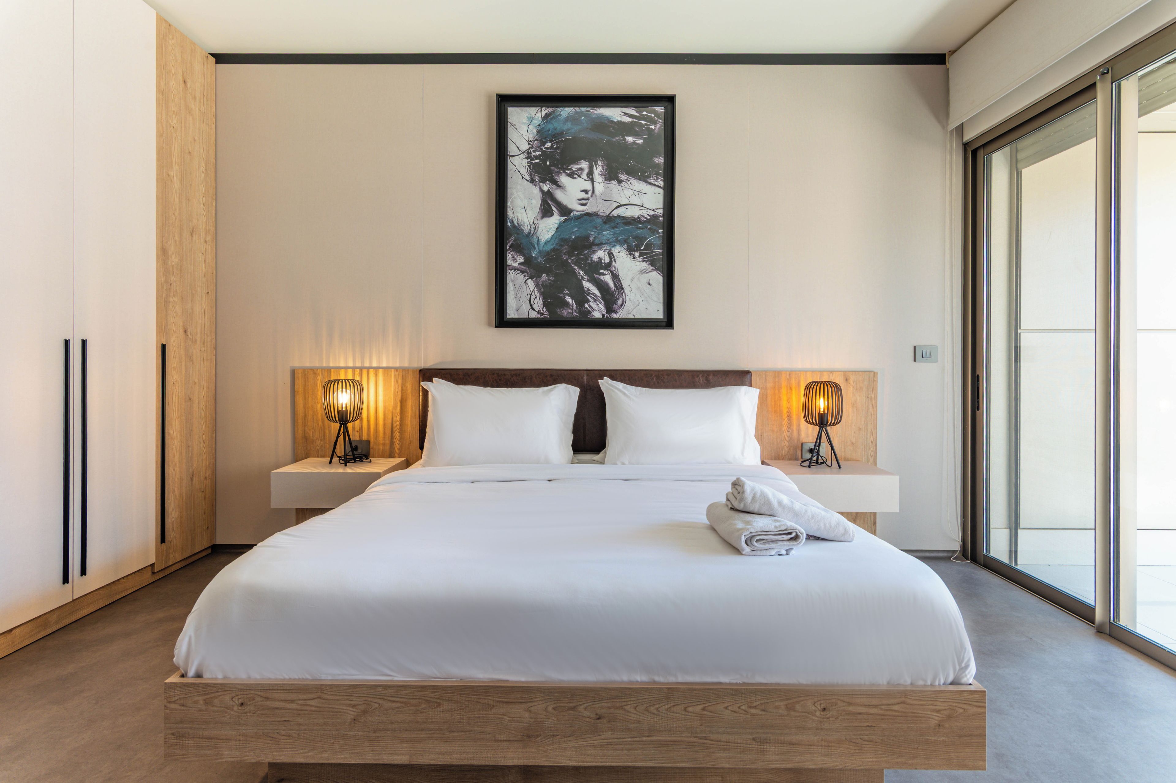 A modern hotel room with a large bed, two bedside lamps, a framed artwork on the wall, and a view through a large window.
