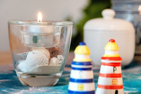 A glass container with a candle, surrounded by seashells and a miniature lighthouse. - Apartment 'Warftoase' (Krummhörn Grimersum)