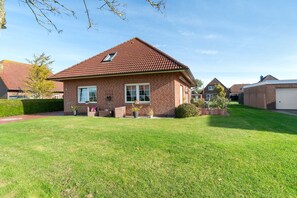 A two-story brick house with a red-tiled roof, a front porch with a bench, and a well-maintained lawn. - Apartment 'Warftoase' (Krummhörn Grimersum)