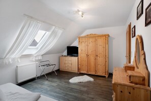 A bedroom with a sloped ceiling, a radiator, a wooden wardrobe, a television, a mirror, and a window with curtains. - Apartment 'Warftoase' (Krummhörn Grimersum)