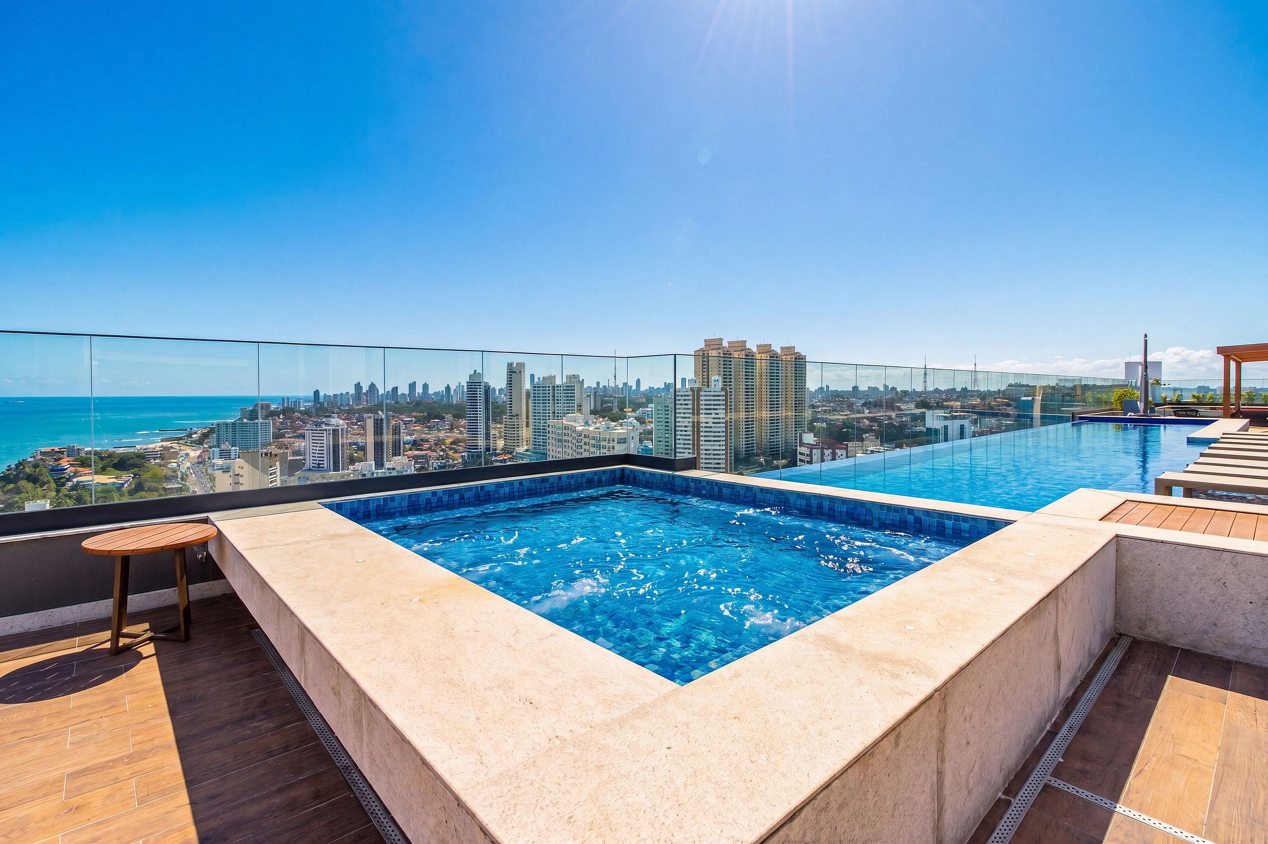 A rooftop infinity pool with a city view, a wooden deck, and a glass balustrade.