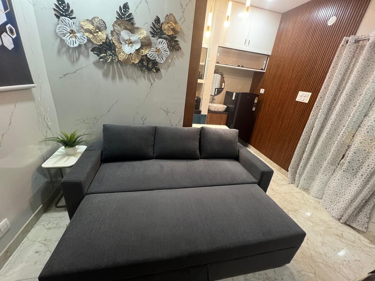 A modern bedroom with a large bed, a decorative wall, a small table with a plant, and a view into the kitchen.