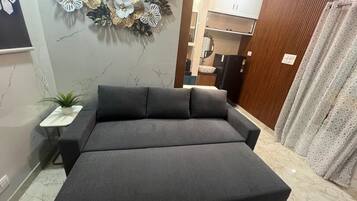 A modern bedroom with a large bed, a decorative wall, a small table with a plant, and a view into the kitchen.