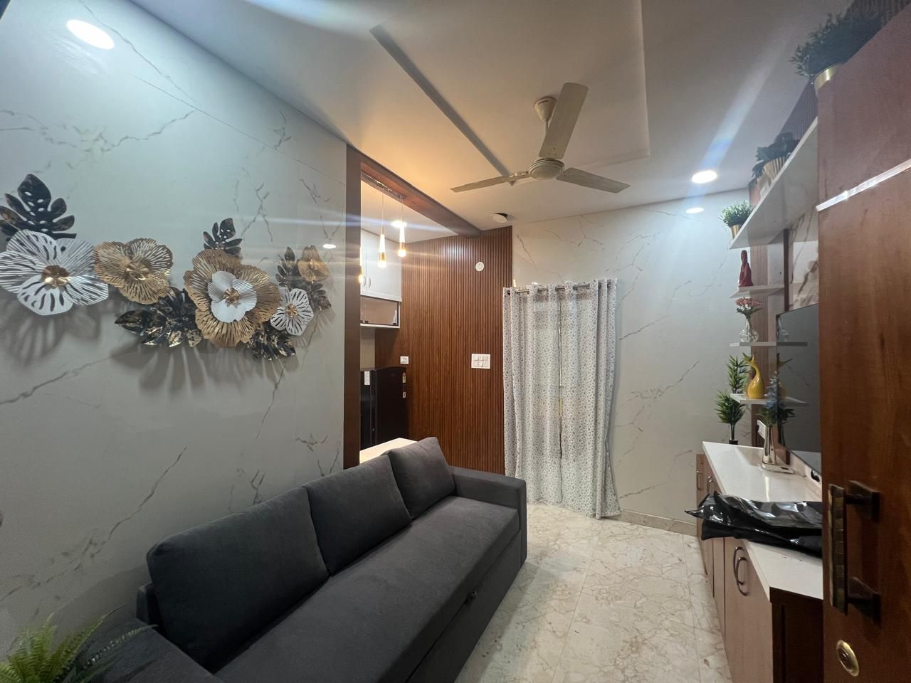 Una sala de estar moderna con un sofá, decoración en la pared, un ventilador de techo y una zona de cocina con armarios y encimera.