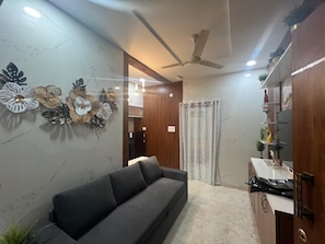 A modern living room with a sofa, decorative wall art, a ceiling fan, and a kitchen area with cabinets and a countertop. - Atlas Home Stays (Hyderabad)