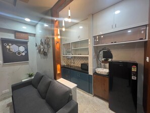 A modern kitchen with dark cabinets, a black refrigerator, and a microwave. There is a dining area with a grey sofa and a small table. - Atlas Home Stays (Hyderabad)