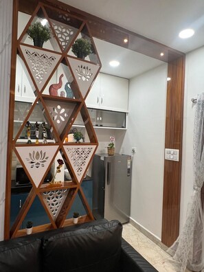 A modern living area with a black sofa, a decorative wooden partition with geometric patterns, a kitchen with white cabinets, and a refrigerator.