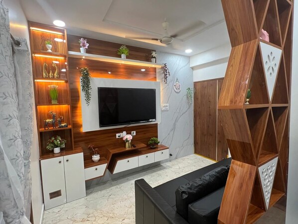 A modern living room with a flat-screen TV, wooden shelving, and decorative plants.