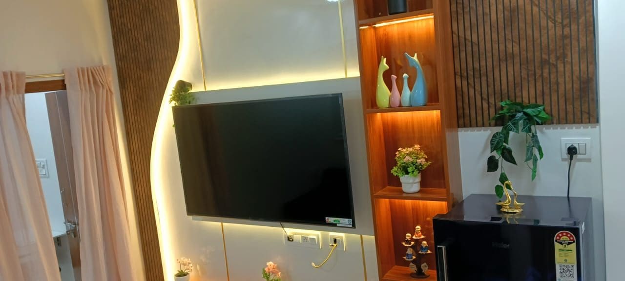 Una sala moderna con un televisor de pantalla plana montado en la pared, estanterías de madera con objetos decorativos y una pequeña planta en maceta.