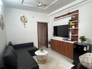 A living room with a black sofa, a white round coffee table, a flat-screen TV, wooden cabinets, and a small plant on the coffee table.