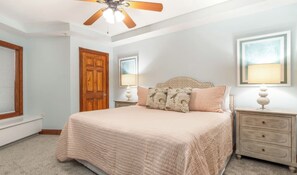 A bedroom with a bed, a wooden door, a ceiling fan, and two bedside tables with lamps. - 235 The Vue  -- New Listing (Tybee Island)