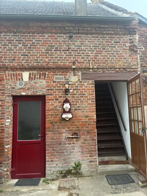 Eine rote Tür mit Fenster, eine Backsteinmauer, ein kleiner Lüftungsschacht, eine Wandleuchte und eine Holztreppe, die zu einer Glastür führt.