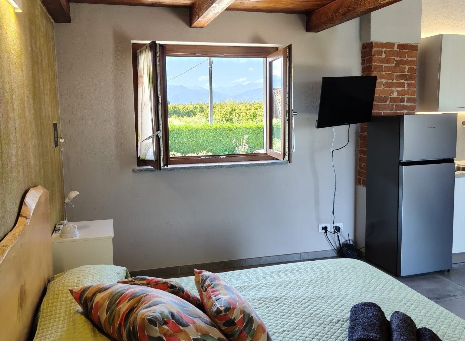 Un dormitorio con una cama, almohadas coloridas, un televisor, un refrigerador y una ventana con vistas al verdor.