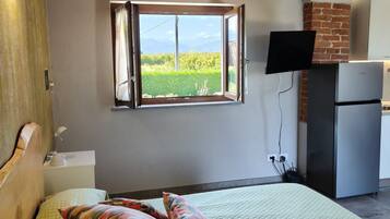 Un dormitorio con una cama, almohadas coloridas, un televisor, un refrigerador y una ventana con vistas al verdor.