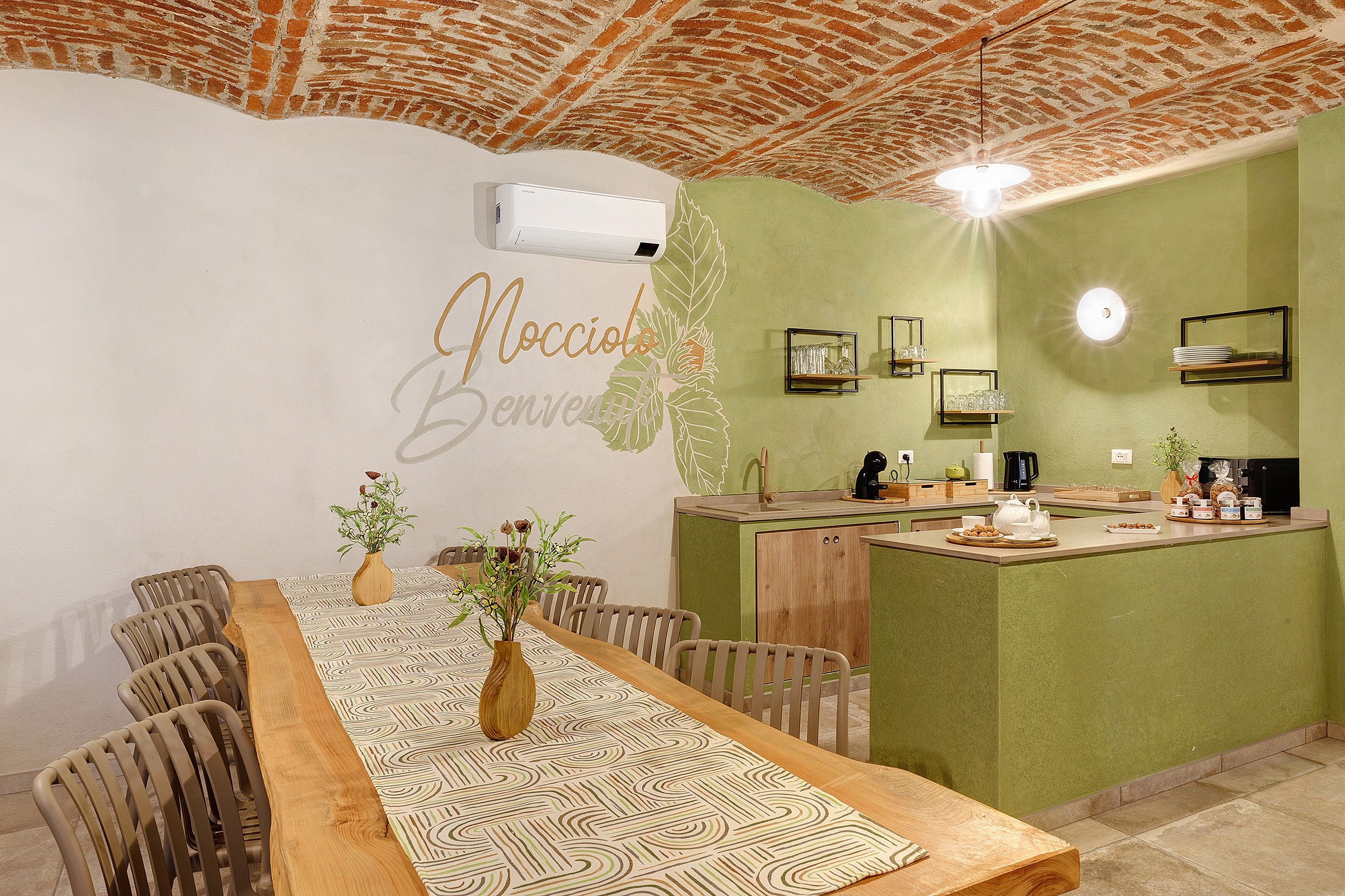 A dining area with a long wooden table, chairs, a vase with flowers, and a kitchen area with a green wall and a brick ceiling.