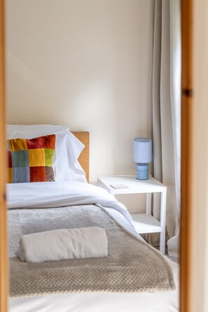 A neatly made bed with a colorful pillow, a bedside table with a lamp, and a window with curtains. - Swift Host | Furtherfield | Sleeps 3 | Countryside | Garden (Reedham)