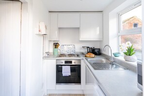 A compact kitchen with white cabinets, a built-in oven, a sink, and a window with a view of a brick building. - Swift Host | Furtherfield | Sleeps 3 | Countryside | Garden (Reedham)