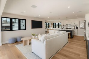 A modern living room with a sofa, a dining area with a kitchen in the background, and large windows. - Brand New Construction | AC | Fire Pit | Garage Parking | Walk to All! (San Diego)