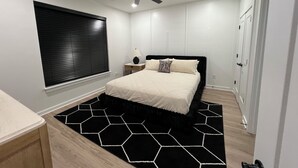 Een slaapkamer met een bed, een plafondventilator, een raam met zonwering, een nachtkastje met een lamp, en een zwart-wit vloerkleed.