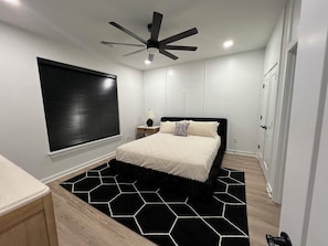 Une chambre à coucher avec un lit, un ventilateur de plafond, une fenêtre avec des stores, une table de chevet avec une lampe et un tapis à motifs noir et blanc.