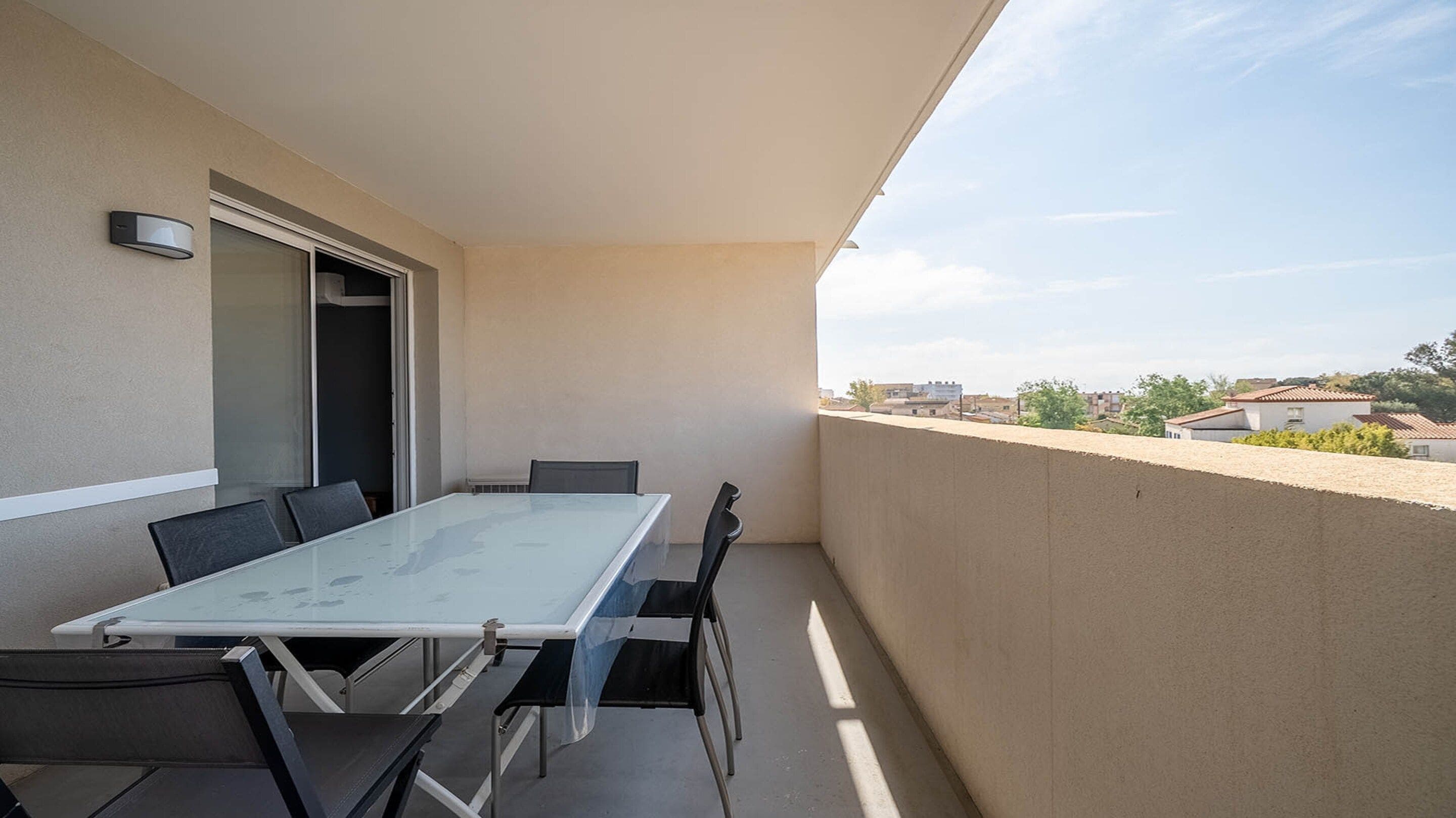 A balcony with a glass table and chairs, a sliding door, and a view of buildings in the distance.