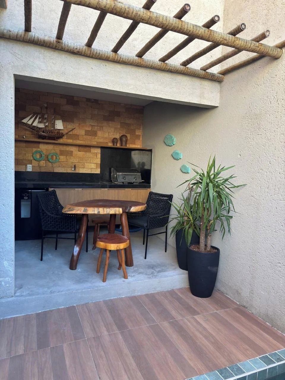 An outdoor patio with a wooden table and chairs, a potted plant, and a brick wall with a grill area.